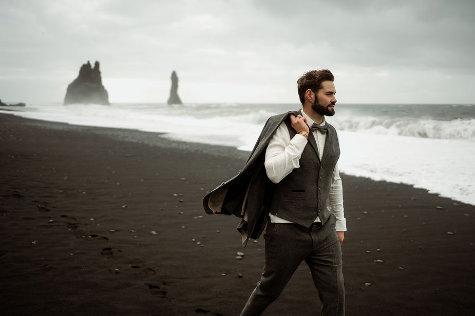 groom walks along black sand beach during iceland honeymoon.