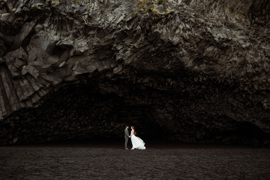 bride and groom stand under rock formations in iceland.