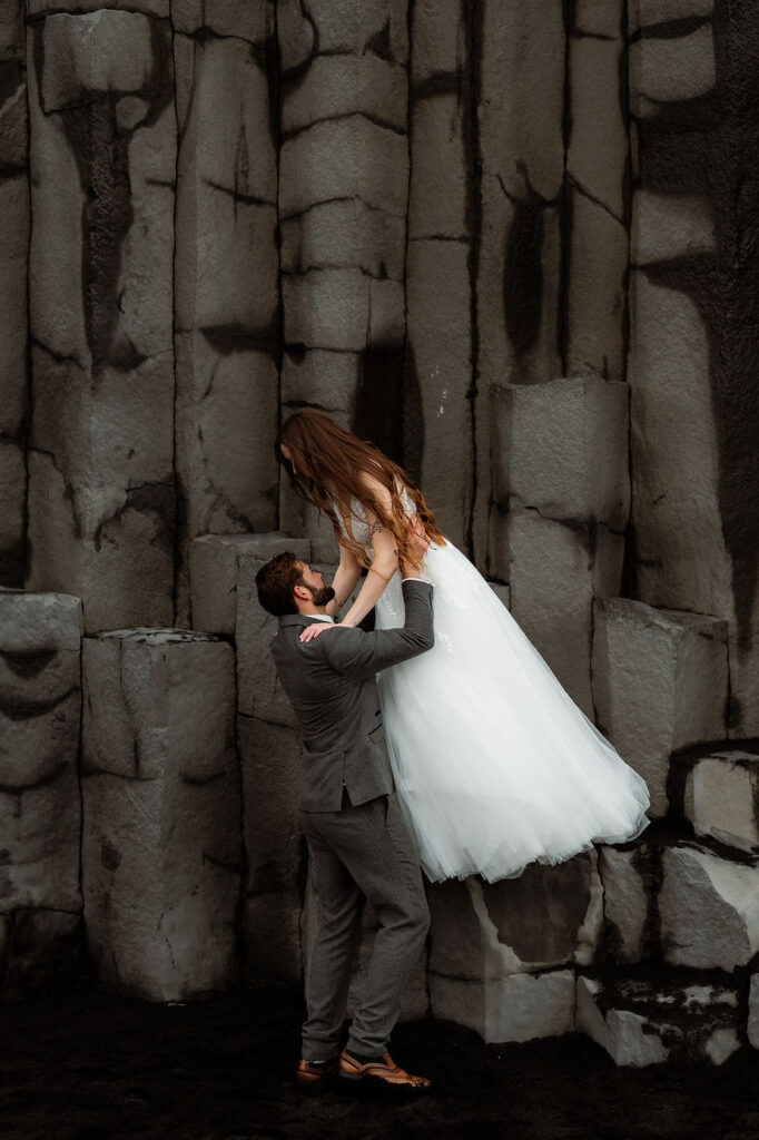 groom lifts bride from rock formations during iceland honeymoon photo shoot.