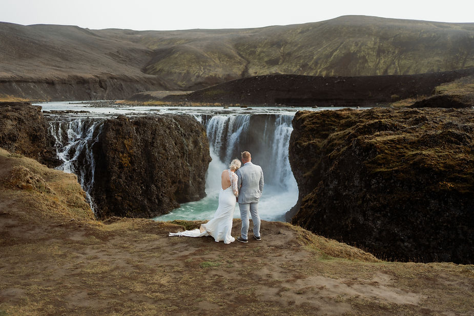 bride and groom look out at waterfall in iceland during wedding day.