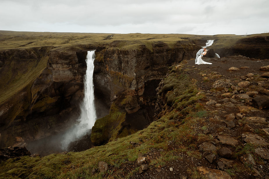 bride and groom kiss on ledge by waterfall in iceland.