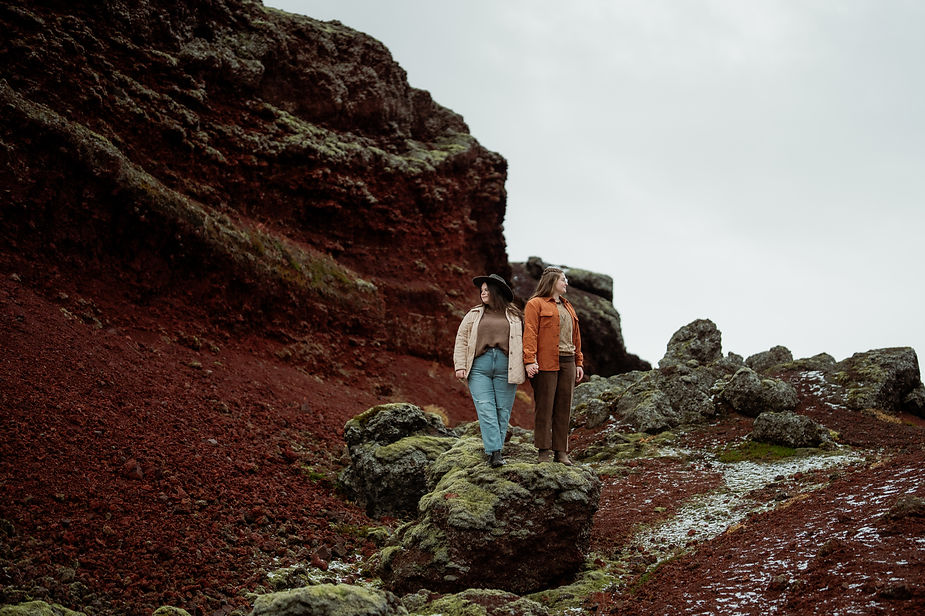 lgbtq+ couple stand on mossy rock and pose for iceland wedding photographer during engagement photos in iceland.