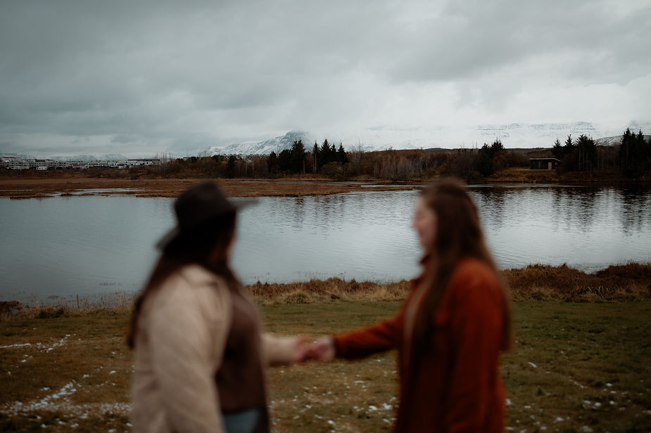 lgbtq+ couple hold hands in front of lake outside of reykjavik during engagement photos.