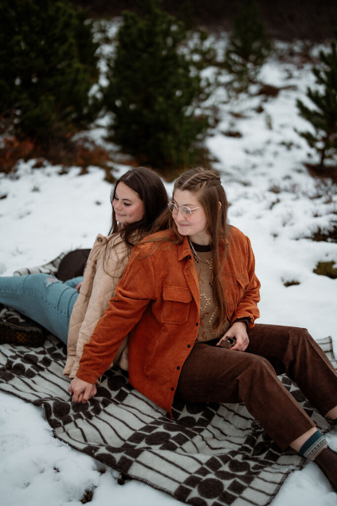 lgbtq+ couple sit on a blanket back to back as one woman holds an engagement ring.