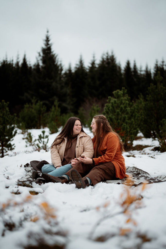 lgbtq+ couple sit on blanket as one woman proposes to the other during their iceland engagement photos.
