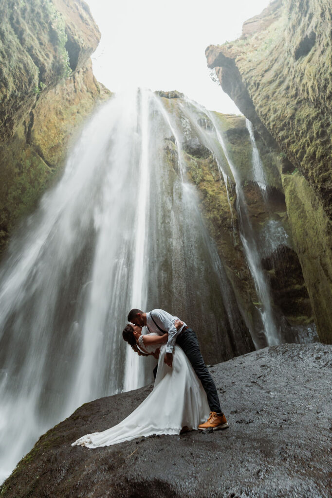 bride and groom stand and kiss on rock under waterfall in Iceland.