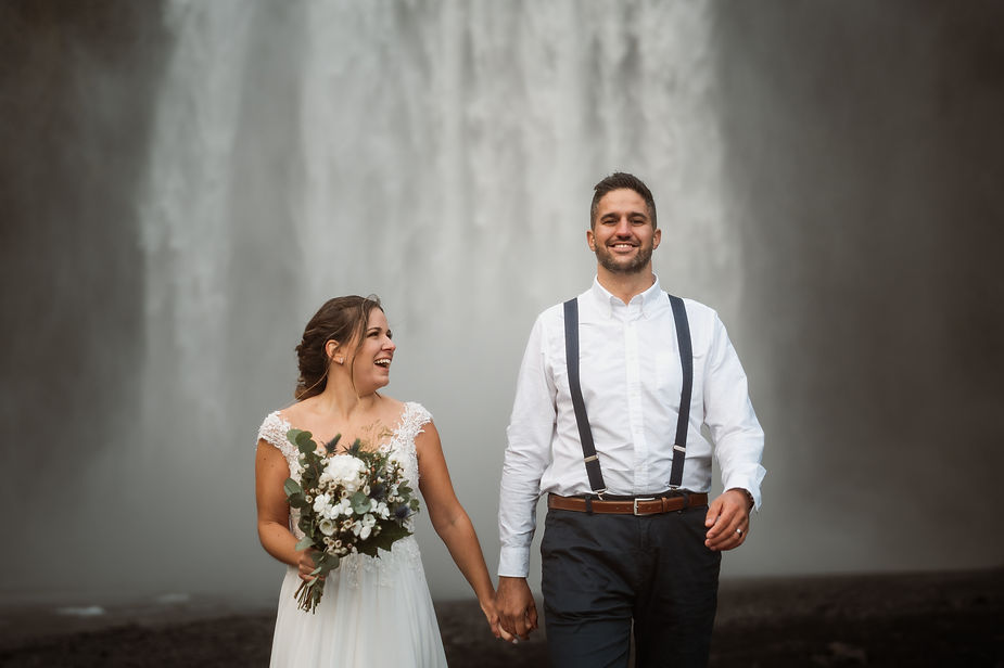 bride and groom walk hand in hand away from iceland waterfall.