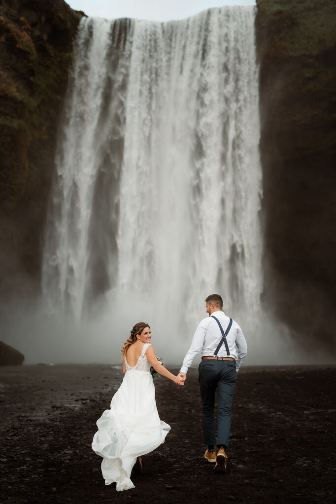bride and groom hold hands and run toward waterfall in iceland.