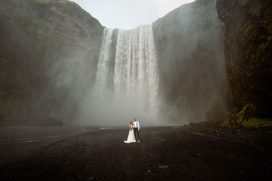 bride and groom stand at base of iceland waterfall.