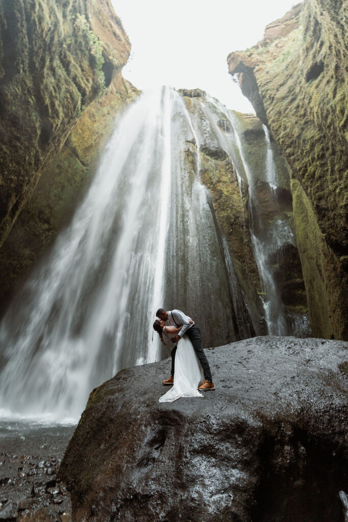 groom dips and kisses bride on a rock at the base of a waterfall in iceland.