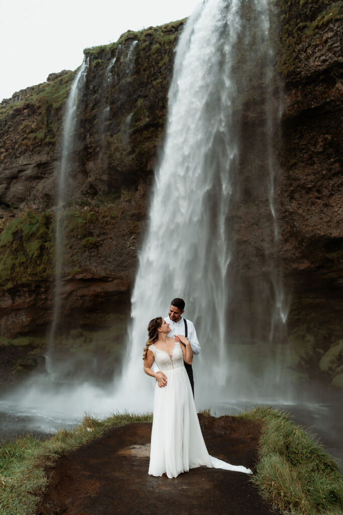 bride and groom stand in front of waterfall in iceland.