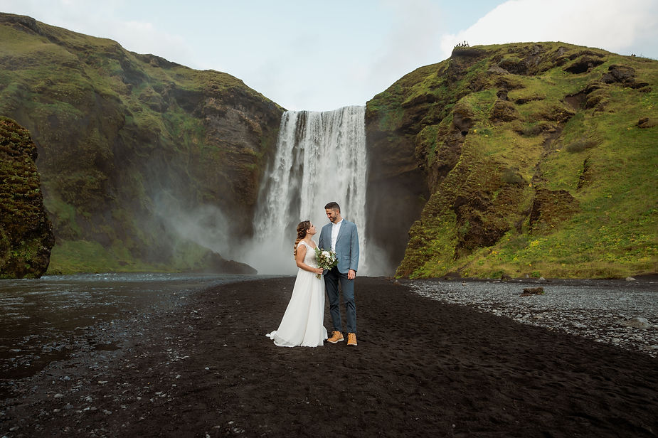 bride and groom stand in front of grassy valley and waterfall in iceland during wedding.