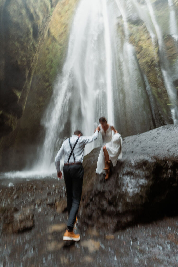 groom helps bride off rock in front of waterfall during iceland elopement.