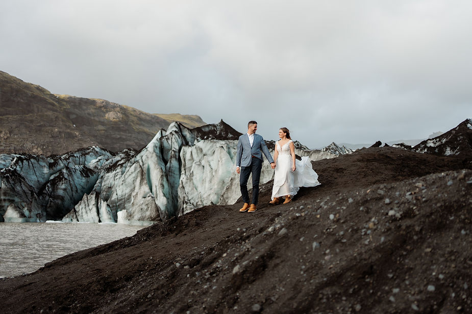 bride and groom walk along sand by glacier in iceland.