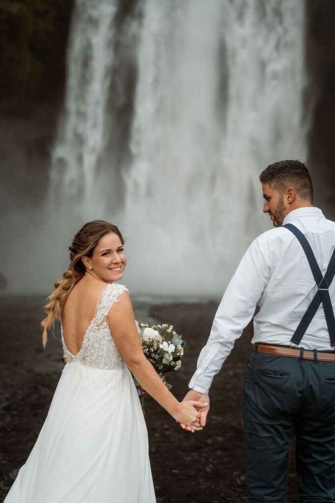 bride and groom hold hands and walk towards waterfall during iceland elopement day.